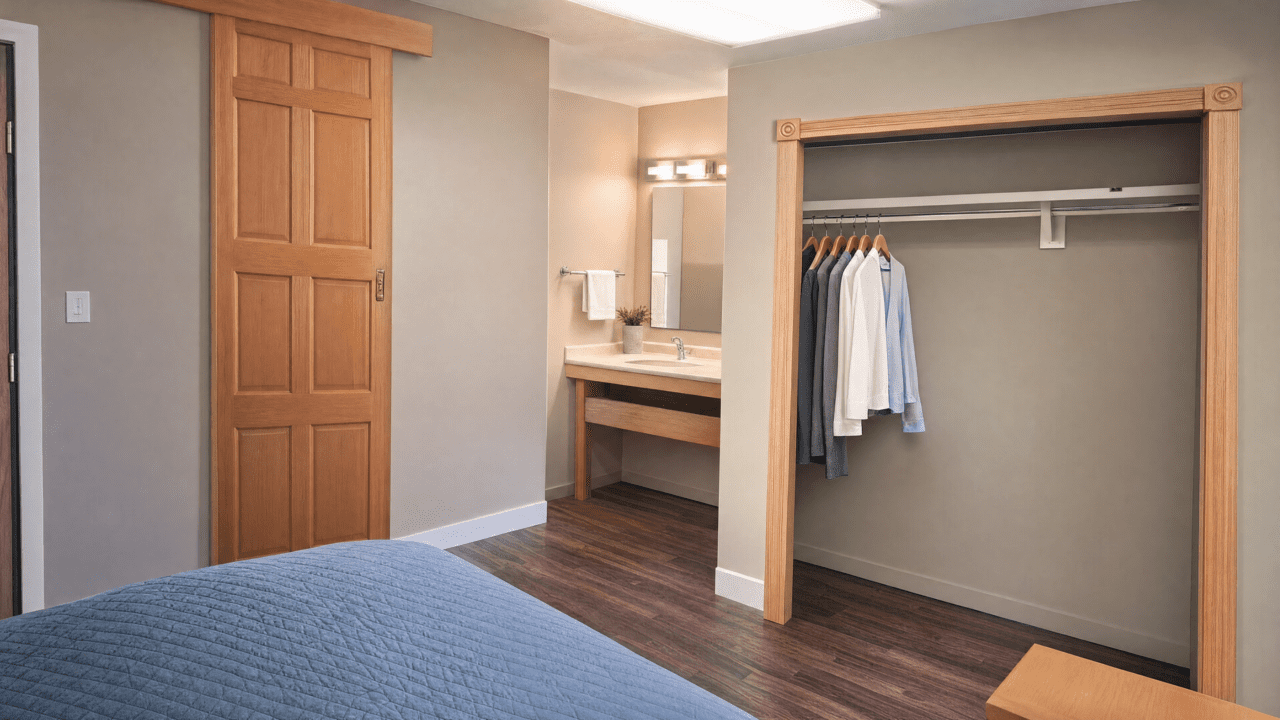 A bedroom featuring a bed with a blue quilt, a wooden sliding closet door with clothes hanging inside, a wall-mounted mirror with a light fixture above it, and a wooden vanity with a sink and a small decorative plant.