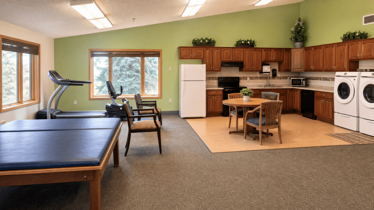 A communal room with large windows, a treadmill and exercise bike on the left, four chairs along the wall, and a kitchenette with wooden cabinets, a white refrigerator, microwave, washing machine and dryer, a small round table with four chairs, and potted plants on top of the cabinets.