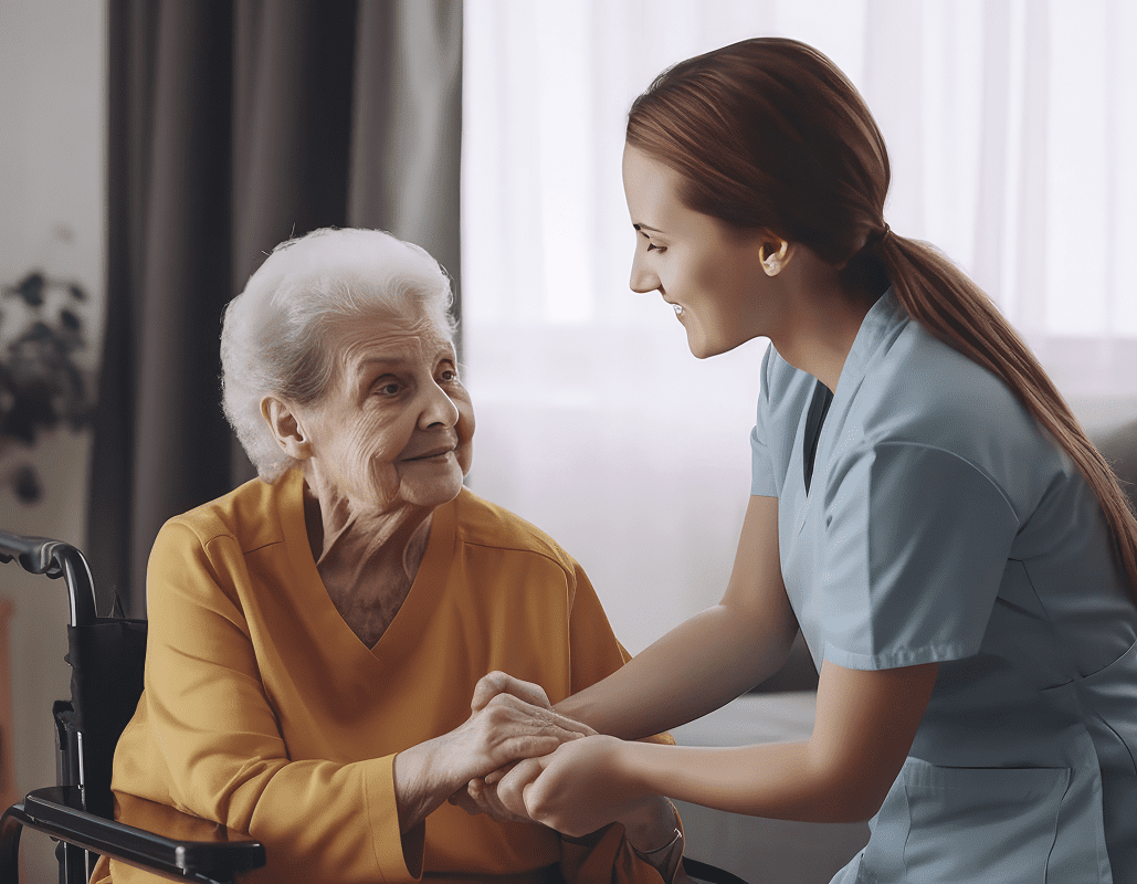 A young female caregiver with red hair smiling warmly as she holds hands with an elderly woman in a wheelchair, in a bright room with curtains in the background.