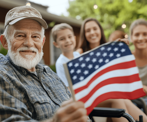 An elderly man with a white beard and gray cap smiling while holding a small American flag, with three happy young adults in the background outdoors.