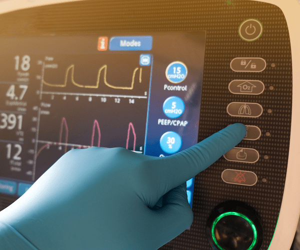 A gloved hand pressing a button labeled with a lung icon on a medical ventilator control panel, which displays various settings and graphs on a screen.