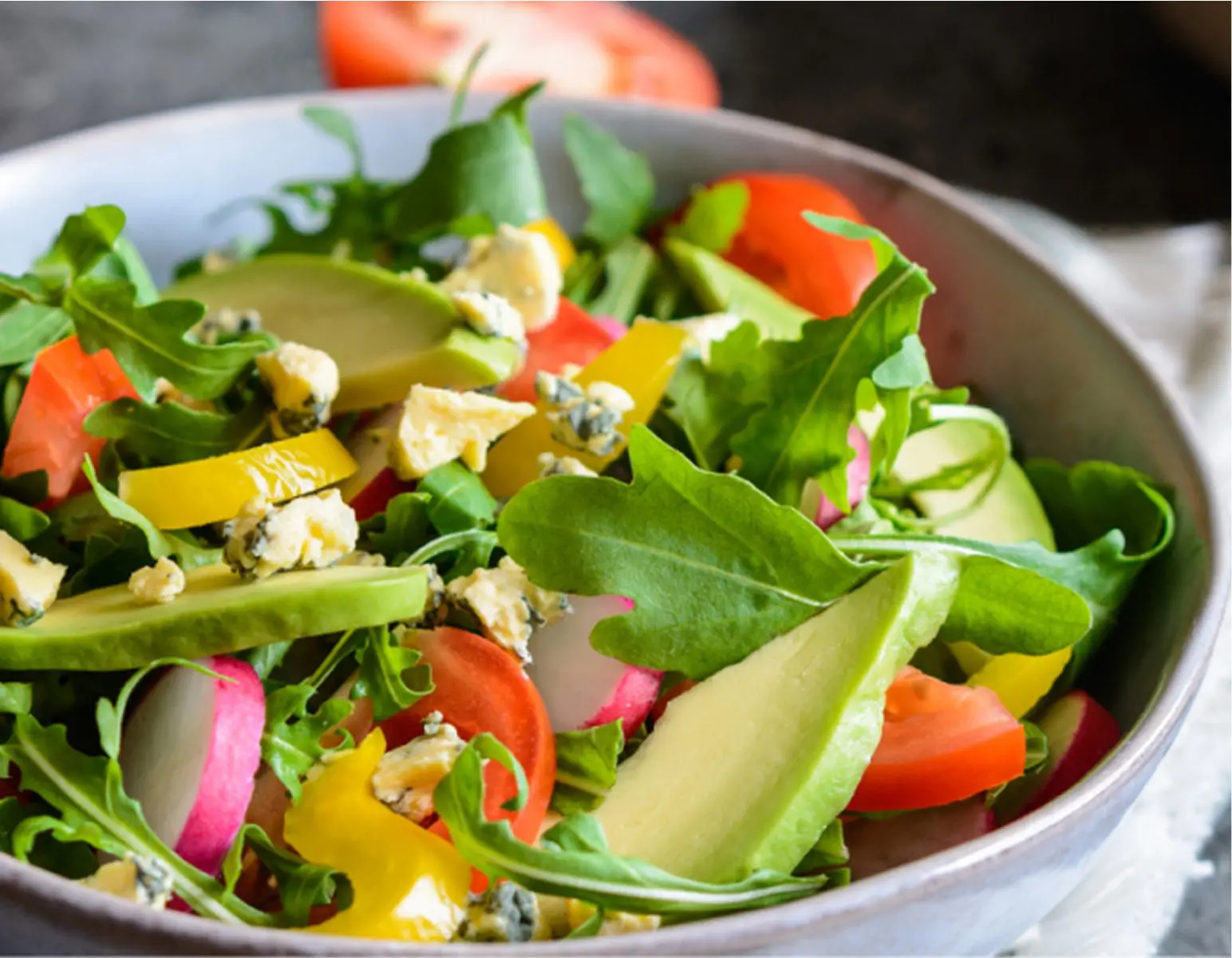 Close-up of a fresh vegetable salad in a bowl, containing arugula, cherry tomatoes, sliced radishes, yellow and green bell peppers, avocado, and blue cheese crumbles.
