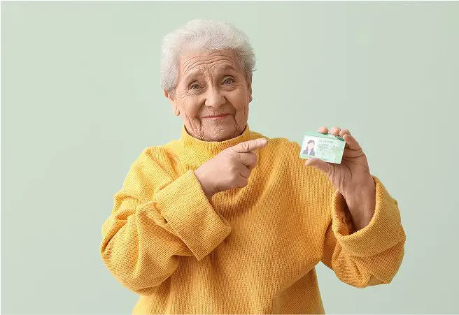 An elderly woman with white hair, wearing a yellow sweater, smiling and pointing at her driver’s license.