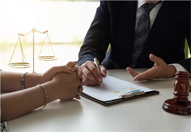 A person in a suit and tie discusses legal documents with another individual, who has their hands clasped on the table. A golden scale symbol representing justice is visible in the background.