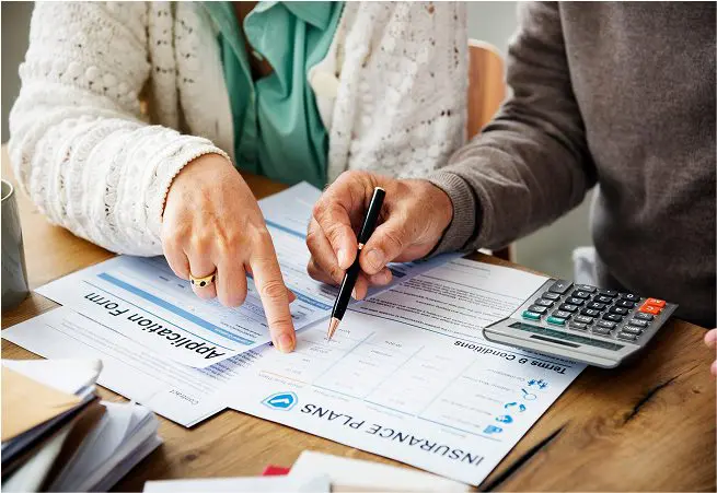 Two people are reviewing insurance documents at a wooden table, with one person pointing to a section on the paper and the other holding a pen. A calculator and stack of papers are also on the table.