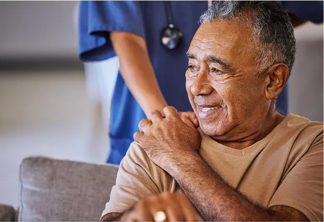 A smiling elderly man sitting on a couch, with a healthcare worker standing behind him. The man has his hand on his shoulder, and the healthcare worker is wearing scrubs with a stethoscope around their neck.