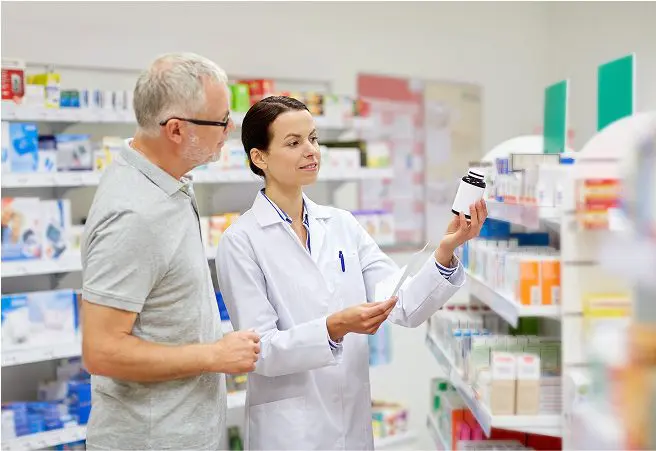 A pharmacy technician in a white coat shows a bottle of medication to an elderly man, both standing in front of pharmacy shelves stocked with various medicines.