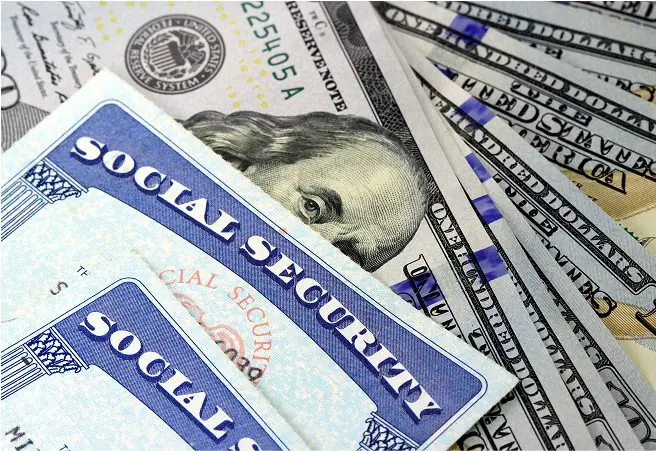 Close-up of a Social Security card placed on top of several U.S. dollar bills, including a $100 bill, with other paper documents partially visible in the background.