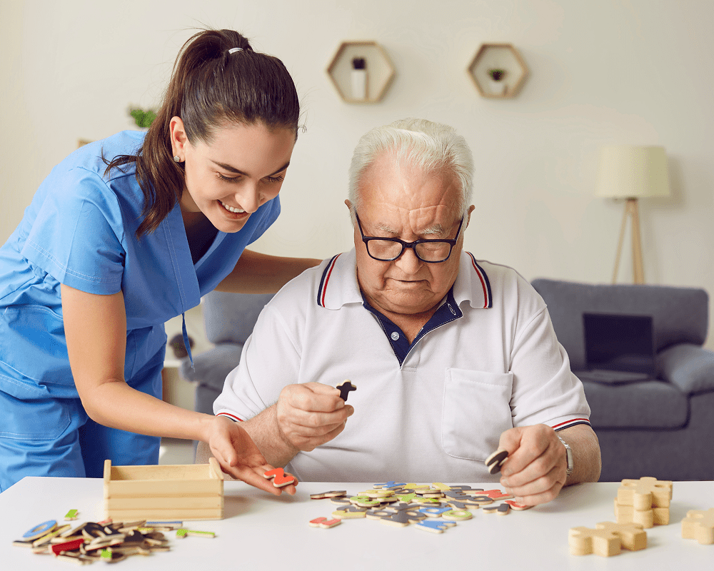 A young female caregiver in blue scrubs assists an elderly man with glasses in playing a game with colorful puzzle pieces at a table. They are in a cozy living room with a gray sofa, a lamp, and decorative wall shelves in the background.